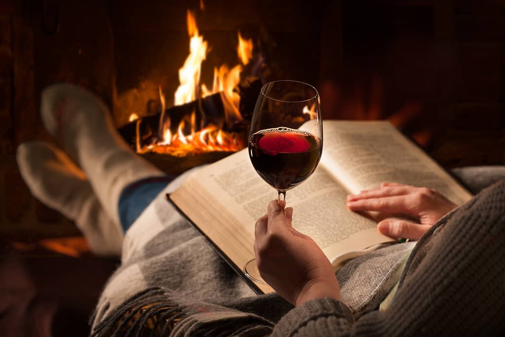 woman reading book with glass of wine in front of fire